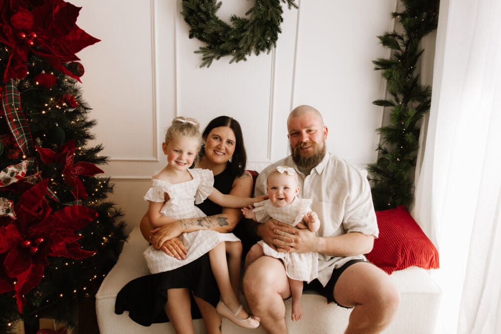 Smiling family of four sitting on a white sofa beside a Christmas tree and wreath during Sweet Moments Photography’s studio Christmas Mini Sessions in Wattle Grove Sydney.